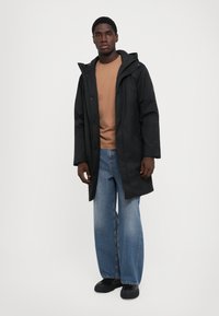 Giacca impermeabile nera con cappuccio, vestibilità rilassata, chiusura a bottoni a scatto, indossata sopra una camicia color sabbia e jeans in denim blu a gamba larga.