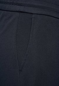 Tessuto di un blu navy scuro con una trama texturizzata, dotato di una tasca laterale e di una cintura liscia. Il materiale appare resistente e leggero.