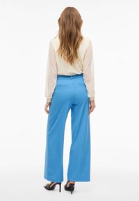 Lehký krémový svetr s rukávy pokrytými puntíky, v kombinaci s modrými kalhotami s vysokým pasem a širokými nohavicemi. Jsou obuty černé vysoké podpatky.