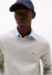 Szürke kötött pulóver kerek nyakkivágással, világoskék csíkos galléros ing fölött. Tengerészkék sapka piros logóval. Simas textúra.