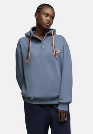 Femme portant un sweat à capuche bleu avec des cordons et des boutons orange, debout avec une expression neutre sur un fond uni.