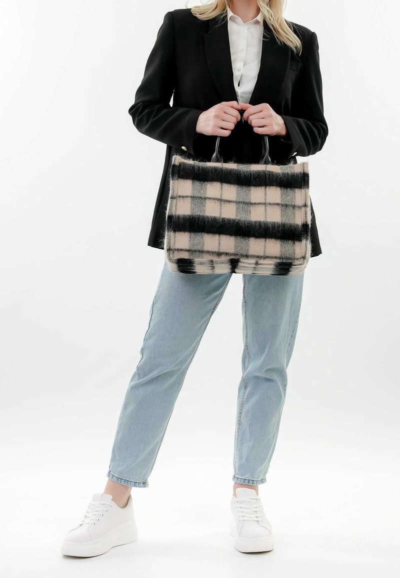 Femme portant un blazer noir, une chemise blanche, un jean bleu clair, des baskets blanches, tenant un grand sac à main beige duveteux à carreaux noirs.