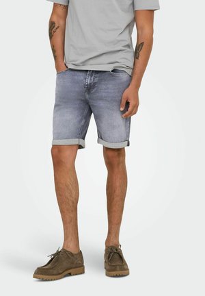 Shorts en denim bleu clair avec des ourlets retroussés, portés avec un t-shirt gris à col rond et des chaussures en daim marron. Les shorts présentent un lavage décoloré.