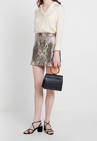 Beige Bluse mit Knöpfen, metallische Schlange-Print-Rock, schwarze Handtasche mit orangefarbenem Griff und schwarze Riemchen-Blockabsatzsandalen.