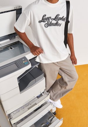 Bílé tričko s nápisem "Low Light Studios" černým písmem, doplněné béžovými kalhotami. Umístěno vedle kopírky se šedými a bílými detaily.