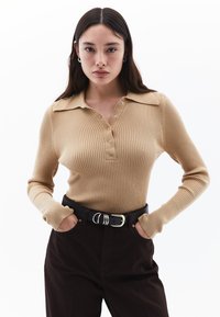 Vaalean beige ribattu pitkähihainen polo, jossa on kaulus, napitus ja se on yhdistetty tummanruskeisiin housuihin ja mustaan vyöhön.