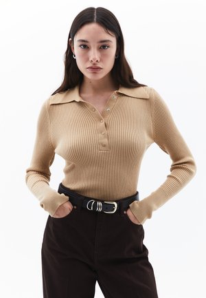 Lichtbeige geribbelde polo met lange mouwen en kraag, voorzien van een knoopsluiting en gedragen met donkerbruine broeken en een zwarte riem.