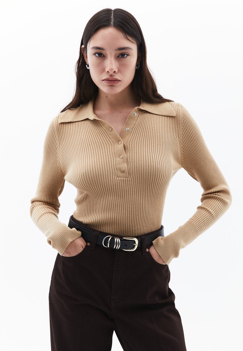 Vaalean beige ribattu pitkähihainen polo, jossa on kaulus, napitus ja se on yhdistetty tummanruskeisiin housuihin ja mustaan vyöhön.