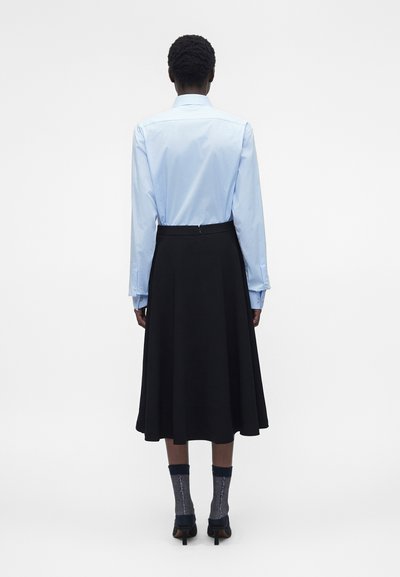 Άτομο που φοράει ανοιχτό μπλε πουκάμισο με κουμπιά, μαύρη midi φούστα, κάλτσες με σχέδια και μαύρες ψηλοτάκουνες, στέκεται γυρισμένο πλάτη σε λευκό φόντο.