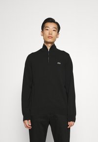 Pulover negru cu fermoar, cu guler înalt, manșete și tiv cu nervuri, având un logo verde Lacoste pe pieptul stâng. Țesătura pare netedă.