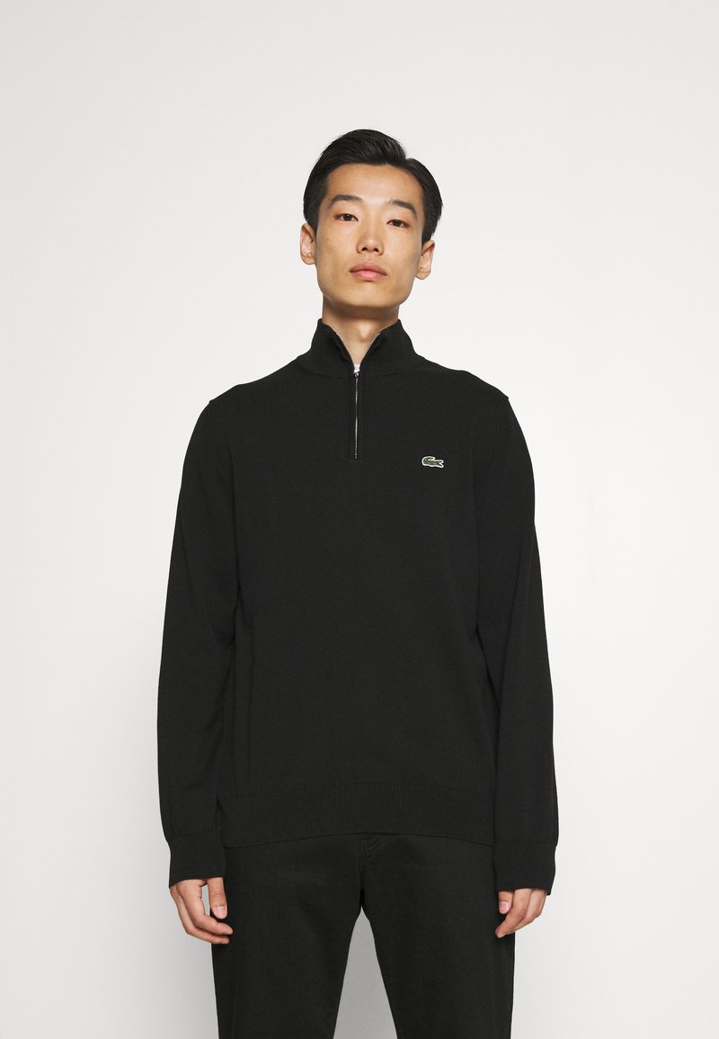 Pulover negru cu fermoar, cu guler înalt, manșete și tiv cu nervuri, având un logo verde Lacoste pe pieptul stâng. Țesătura pare netedă.