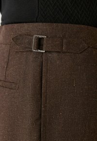 Pantalons en tissu marron avec une finition texturée, présentant une boucle en métal argenté, un devant plat et des poches latérales.
