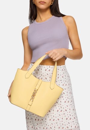 Vrouw met een lavendelkleurige mouwloze top en een bloemenrok, die een grote gele handtas met gouden kettingdetail vasthoudt, voor een witte achtergrond.