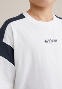 Balta trumpomis rankovėmis marškinėliai su tamsiai mėlynomis pečių detalėmis ir „SALTYDOG“ logotipu tamsiai mėlyna spalva ant priekio. Medvilnės audinys, laisvas kirpimas.
