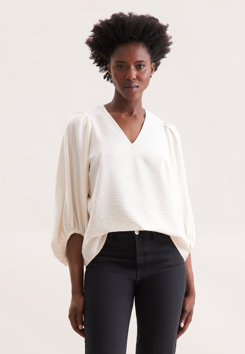 Hvid V-hals bluse med puffede ærmer, struktureret stof. Parret med højhævede sorte jeans med lukning foran.