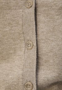 Beige cardigan laget av mykt stoff, med ribbestrikk langs kantene og fem runde knapper foran.