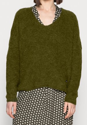 Olivgrön, löst sittande tröja i mjukt, texturerat material. Har en djup V-ringning och långa ärmar, bärs över en klänning med prickar.