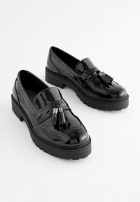 Mocasines de charol negro con suela plana, que presentan borlas decorativas y detalles de brogue a lo largo de los bordes.