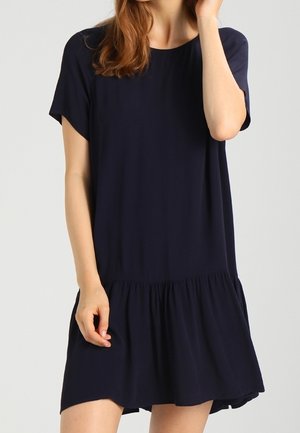 Kobieta w luźnej, granatowej sukience z krótkim rękawem i marszczonym falbanowym dołem, stojąca na jasnym, jednolitym tle.