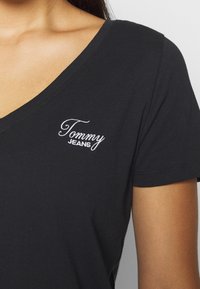 Detailní záběr na osobu v černém tričku s V-výstřihem, na levé straně hrudi vyšívané bílé "Tommy Jeans".