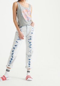 Canotta grigia senza maniche con stampa grafica colorata, abbinata a jeans azzurri a tie-dye con risvolto e sandali bianchi slip-on con dettagli rossi.