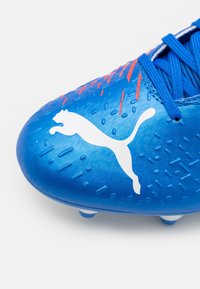 Scarpino da calcio blu con superficie testurizzata, logo bianco e dettagli rossi. Presenta una punta arrotondata, lacci visibili e numerosi tacchetti sulla suola.