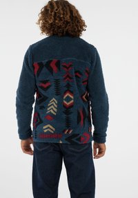 Person mit lockigem Haar, der eine blaue Fleecejacke mit roten, schwarzen und beigen geometrischen Mustern trägt, kombiniert mit dunkelblauen Jeans, von hinten gesehen.