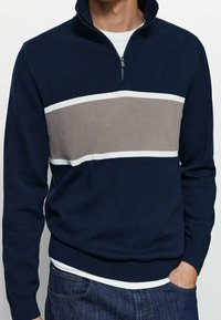 Homme portant un pull bleu marine avec des rayures horizontales grises et blanches, col à fermeture éclair, et un jean bleu, main dans la poche.