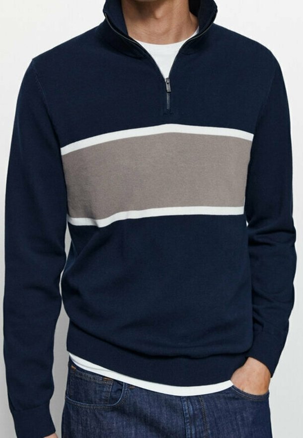Homme portant un pull bleu marine avec des rayures horizontales grises et blanches, col à fermeture éclair, et un jean bleu, main dans la poche.