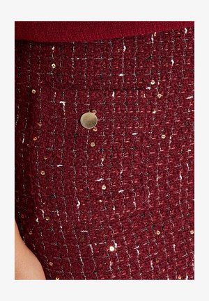 Falda burdeos texturizada hecha de tela tejida con acentos metálicos y lentejuelas doradas. Presenta un botón circular dorado en el costado.