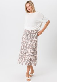 Lichte, crèmekleurige trui gecombineerd met een gemarmerde bruine rok met geometrische ontwerpen en een zijnaad, gedragen met witte sandalen.