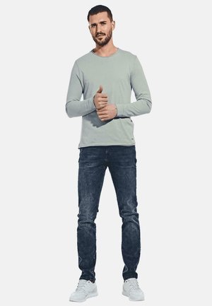 Light gray long-sleeve shirt with a round neckline, paired with dark blue jeans and white athletic shoes. Model stands with a thumbs-up gesture.