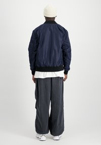 Marine bomberjakke med ribbede krave, parret med løse grå bukser og hvide sneakers, set bagfra.