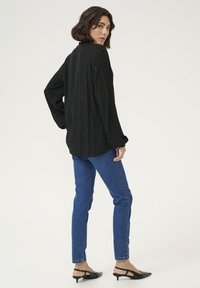 Schwarzes Langarmoberteil mit vertikalen metallischen Streifen, kombiniert mit blauen Slim-Fit-Jeans und schwarzen Slingback-Absätzen, von hinten betrachtet.