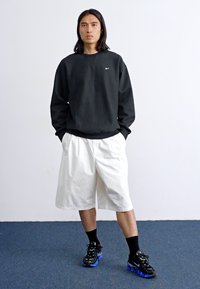 En person med langt hår iført en sort sweatshirt, hvide shorts og sorte sneakers står op ad en ensfarvet hvid væg.