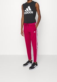 Schwarzes ärmelloses Sporttop mit weißem "adidas"-Logo, kombiniert mit enganliegenden rosa Hosen mit weißen Seitenstreifen und schwarzen Sportschuhen.