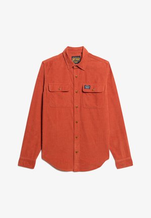 Camisa de pana en naranja quemado, con un frente de botones, dos bolsillos en el pecho y una etiqueta en el lado izquierdo con detalles del logo.