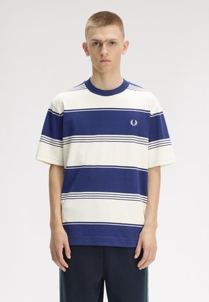Giovane uomo in piedi, frontale, indossa una camicia a maniche corte a righe larghe blu e bianca con pantaloni blu scuro su sfondo semplice.