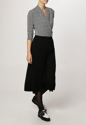 Vrouw die een zwart-wit pied-de-poule top draagt, een zwarte geplooide midirok, zwarte panty's en zwart-witte loafers terwijl ze staat.