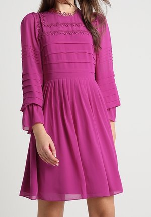 Femme portant une robe fuchsia mi-longue à manches longues, avec une jupe plissée, des manches superposées et des détails en dentelle florale sur la poitrine et le décolleté.