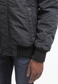 Giubbotto bomber in tessuto trapuntato nero con tasca zip, polsini a costine e orlo testurizzato. Presenta bottoni tondi per la chiusura e una finitura liscia.