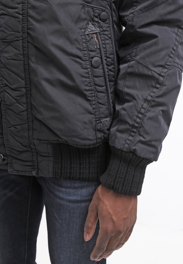 Giubbotto bomber in tessuto trapuntato nero con tasca zip, polsini a costine e orlo testurizzato. Presenta bottoni tondi per la chiusura e una finitura liscia.