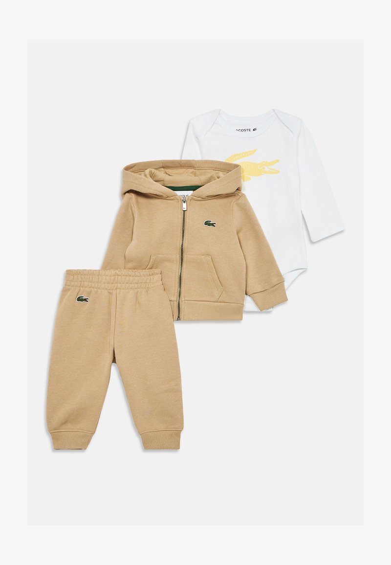 Hoodie bege com fecho, com bolsos, calças de moletom a condizer e um bodysuit branco de manga longa com um logótipo de crocodilo amarelo. Material em algodão.