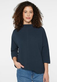 Marineblaues, strukturiertes Oberteil mit Dreiviertelärmeln und hohem Ausschnitt, kombiniert mit hellblauen Jeans aus Denim in lockerer Passform.