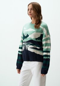 Pull en tricot mettant en avant un paysage de montagne en vert, bleu marine et blanc. Le design inclut des rayures texturées et une coupe décontractée.