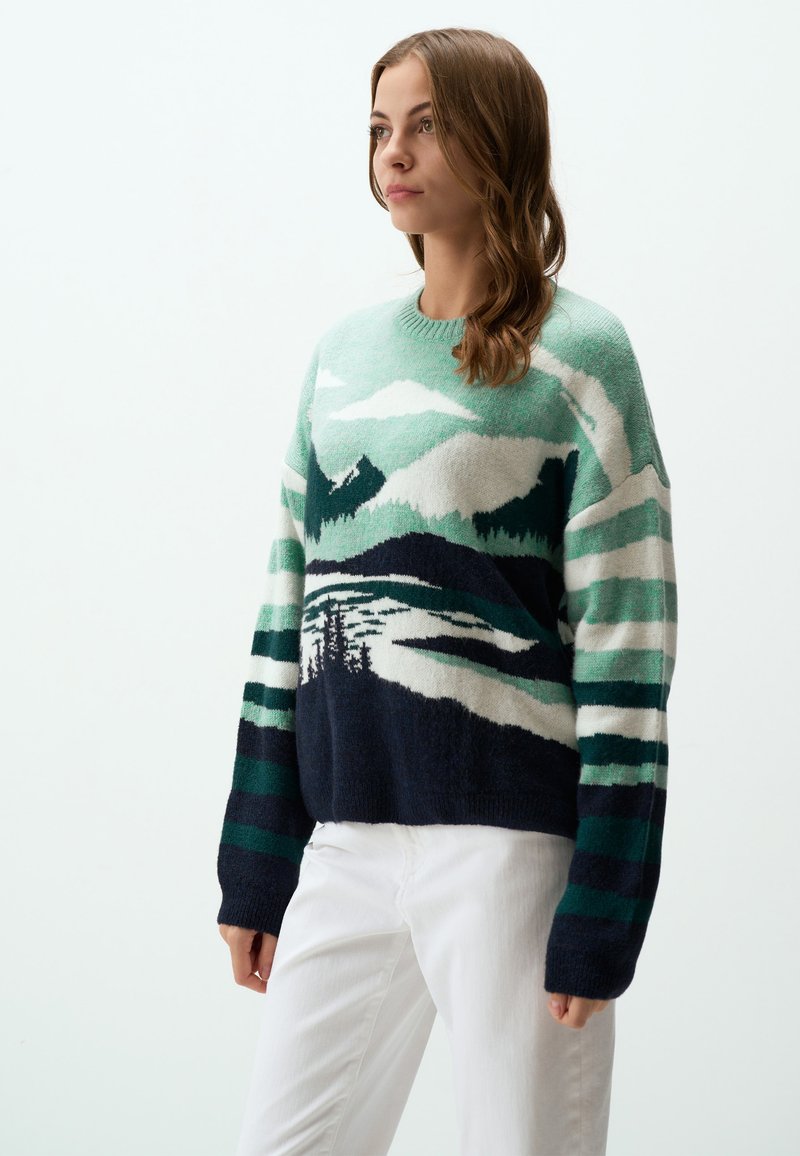 Pull en tricot mettant en avant un paysage de montagne en vert, bleu marine et blanc. Le design inclut des rayures texturées et une coupe décontractée.
