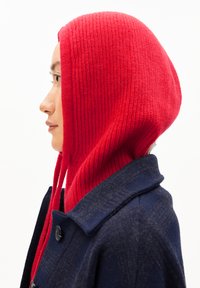 Rote, gerippte Strickhaube mit langen Bändern, getragen über einem dunkelblauen Mantel. Die Haube hat eine enge Passform, die die Textur und den Farbkontrast betont.