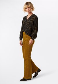 Blusa nera con fantasia, maniche lunghe e scollo a V, abbinata a pantaloni a zampa di elefante in velluto a coste senape e scarpe nere lucide.