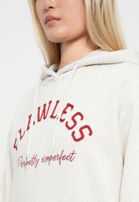 Ανοιχτό γκρι φούτερ από απαλό ύφασμα, με κόκκινα κεντημένα γράμματα "ΑΤΕΛΕΙΩΤΟΣ" και "Τέλεια ατελής." Περιλαμβάνει λευκές κορδόνες.