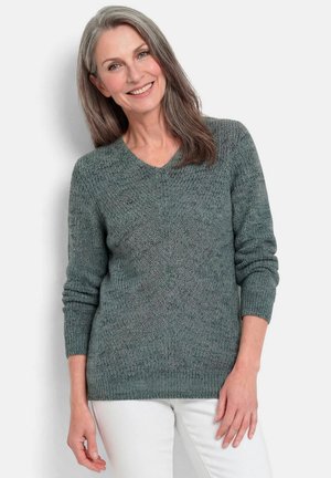 Vrouw van middelbare leeftijd met lang grijs haar draagt een textuurgebreide groene trui en witte broek, glimlachend tegen een effen achtergrond.